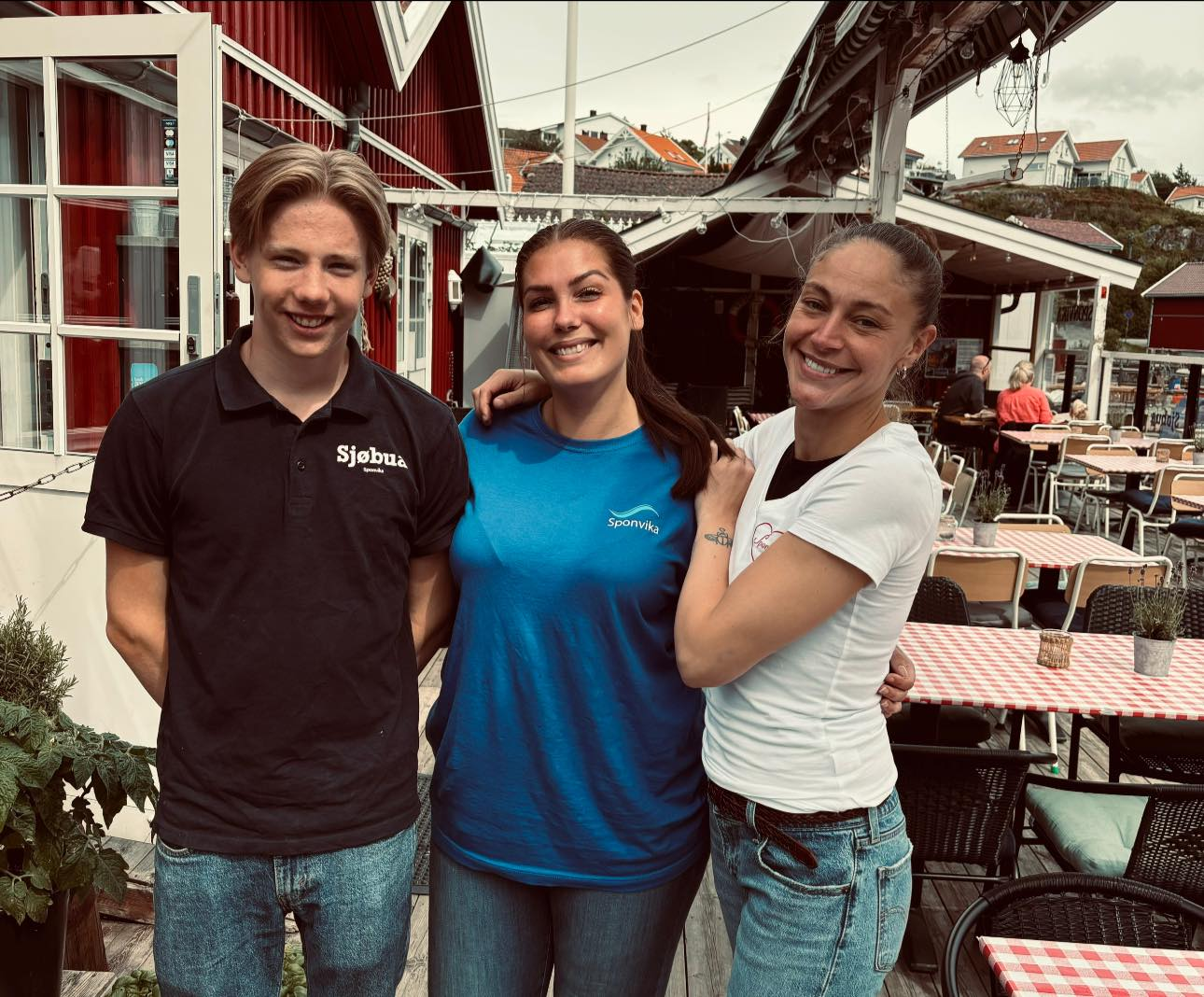 Tre unge medarbeidere fra Sjøbua i Sponvika smiler på terrassen – blide verter klare til å ta imot gjester ved Ringdalsfjorden